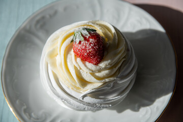 Close-up top view of meringue pavlova dessert with strawberry on saucer standing on colourful wooden table in cafe in a sunny day. Light and shadow. Soft focus. Sweet food theme.