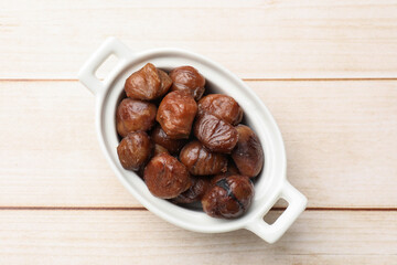 Roasted edible sweet chestnuts in dish on light wooden table, top view