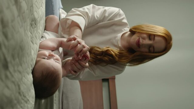 Woman playing with baby on bed. Mother red-haired caucasian happy cheerful woman mommy holds baby's child's hands, plays, develops child's kid's motor skills, helps to learn to move. Inverted frame.