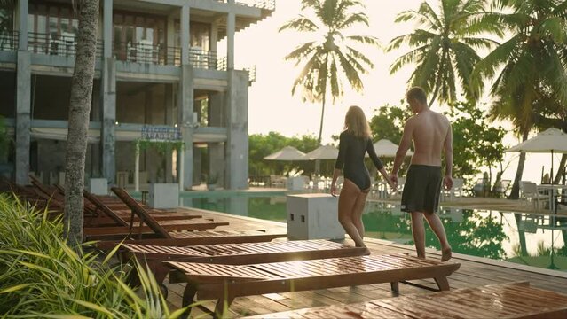 Young couple in swimsuits walking along swimming pool of luxury resort hotel on ocean shore. Cheerful girl and guy holding hands near swimming pool. Happy two people relax on poolside together.