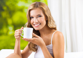 Portrait  image of happy smiling woman drinking coffee sitting near window, at home bedroom. Cheerful amazed blond girl drink indoors.