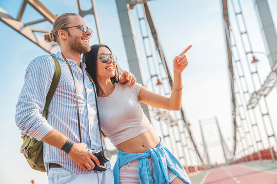 Happy explorer couple standing outdoors. Man talking to woman with his arm around her shoulders and she pointing towards. Traveling and spending time together