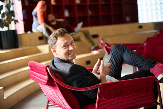 Handsome man smiling sitting in cozy red armchair and surfing internet