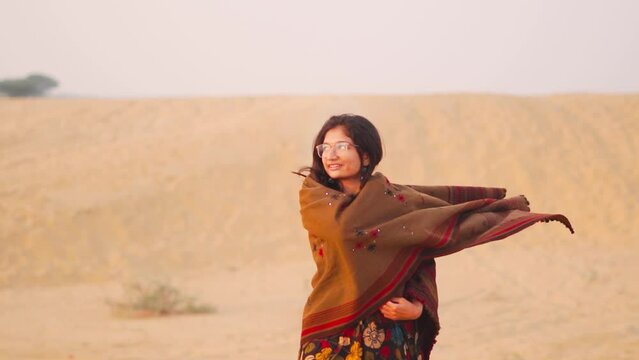 Beautiful Indian woman wearing shawl standing in Thar desert at Sam Sand Dunes in Rajasthan, India. Indian female tourist in desert. Young woman enjoying travel holidays in dessert. 