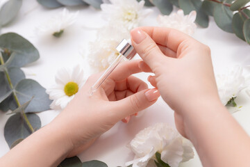 Organic oil for health of nail and skincare concept. Women hands hold pipette with cosmetic serum on white background with flowers, top view