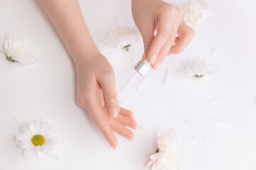 Obraz na płótnie Canvas Women hands hold pipette with cosmetic serum on white background with flowers, top view. Organic oil for health of nail and skincare concept