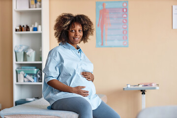 Medium full shot of smiling pregnant African American woman looking at camera and putting hands on...