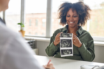 Medium shot of cheerful pregnant Black woman looking at unborn baby sonogram image during...