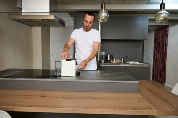 Young adult man preparing his morning espresso using a capsule coffee machine for home use in the minimalist home kitchen