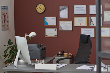 Interior of empty doctor office in hospital with computer desk and framed certificates on wall