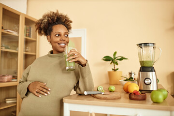 Waist up shot of cheerful African American expectant mother in kitchen holding glass of fresh...