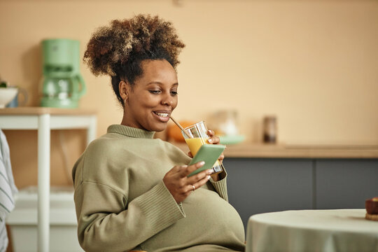 Medium shot of laughing pregnant Black woman with big belly looking at smartphone and holding glass of orange juice with straw sitting at kitchen table