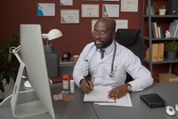 Black doctor filling medical form sitting at his desk while having video call with his patient
