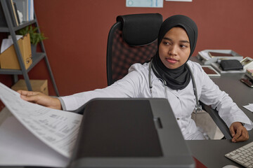 Young muslim doctor making copy of medical document sitting at her desk in office