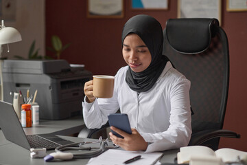 Smiling female doctor in hijab using her smartphone while having coffee break at her workplace