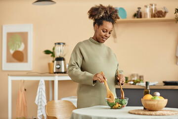 Fototapeta na wymiar Waist up shot of smiling pregnant African American woman mixing fresh salad in bowl standing at kitchen table