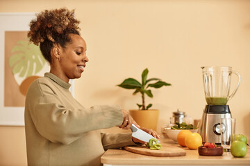 Side view of happy pregnant Black woman with knife cutting kiwi fruit on wooden chopping board in...