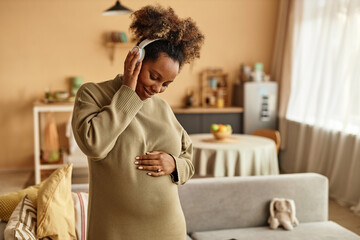 Medium shot of smiling African American expectant mother in headphones listening to calming music...