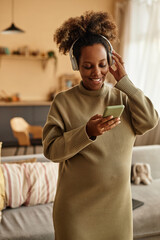 Vertical medium long shot of cheerful African American expectant mother in wireless headphones watching funny videos on smartphone at home