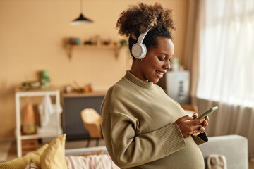 Medium shot of smiling pregnant Black woman at home wearing headphones holding smartphone and...