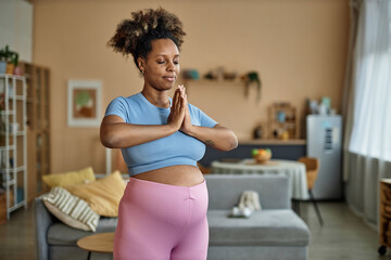 Fototapeta na wymiar Medium full shot of calm pregnant African American woman dressed in sports clothing closing eyes while doing standing meditation in living room with palms folded
