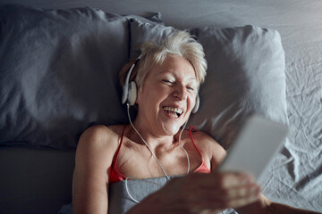 Happy senior woman listening and enjoying relaxing music in bed
