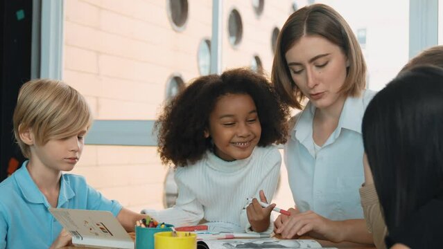 Professional caucasian teacher telling story to diverse student while sitting at table with storybook and colored book. Smart learner listening story while colored picture from instructor. Erudition.