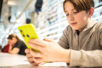 An 11-year-old boy holds a yellow, green smartphone in his hands, looks at the phone screen, sits at a table in a bright public library.
