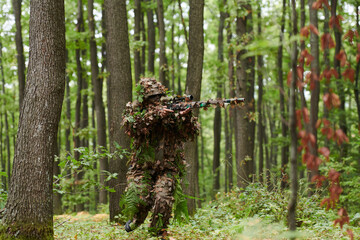 A highly skilled elite sniper, camouflaged in the dense forest, stealthily maneuvers through...