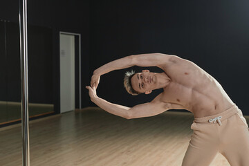 Medium long shot of young Caucasian male pole dancer doing stretching exercise standing in studio