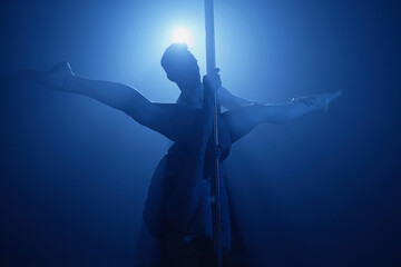 Silhouette of couple performing sensual pole dance together with female hanging upside down doing split and male partner assisting in blue spotlight