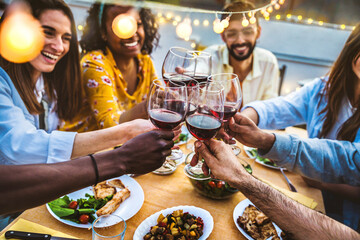 People toasting red wine glasses on rooftop dinner party - Happy friends eating meat and drinking wineglass at restaurant patio - Food and beverage lifestyle concept with guys and girls dining outdoor