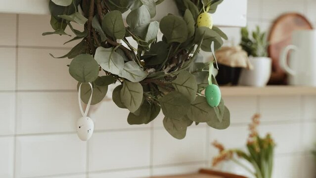 Close-up of the bottom of wreath decorated with decorative colored eggs on ribbons. Hanging spring reusable decoration in home kitchen made from artificial eucalyptus branches, no people.