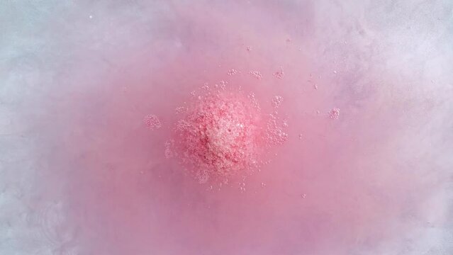 Pink bath bomb ball in blue background dissolves in water with white foam bubbles. Cosmetic product natural and skin care. Clear water with reflections sunlight and shadows. High quality 4k footage