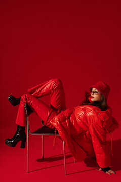 Fashionable confident woman wearing trendy oversized red puffer down jacket, sunglasses, bucket hat, faux leather pants, platform boots, posing on red background. Full-length studio fashion portrait