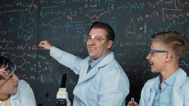 Creative teacher pointing chemistry at blackboard while talking to high school boy at table with microscope and test tube with colored liquid. Young student wearing lab coat in STEM class. Edification