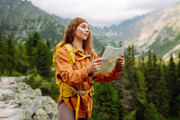 Naklejka premium Young woman traveler with a yellow backpack, holds a map, explores hiking trails in the mountains. The concept of travel, vacation. Hiking and adventure concept