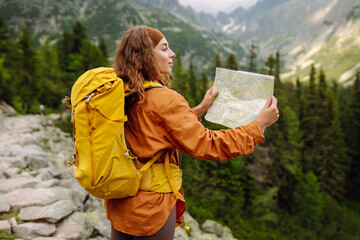 Young woman traveler with a yellow backpack, holds a map, explores hiking trails in the mountains....