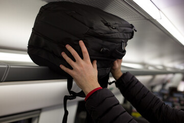 Male hands put a black backpack on the top berth in a train while traveling. Storage space above the seats of public transport under the ceiling - obrazy, fototapety, plakaty
