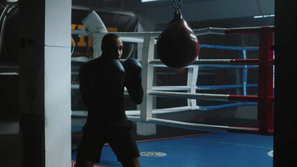Obraz premium Confident African American fighter hits punching bag while training in dark boxing gym. Athletic man in boxing gloves exercises before fighting competition. Physical activity and intensive workout.