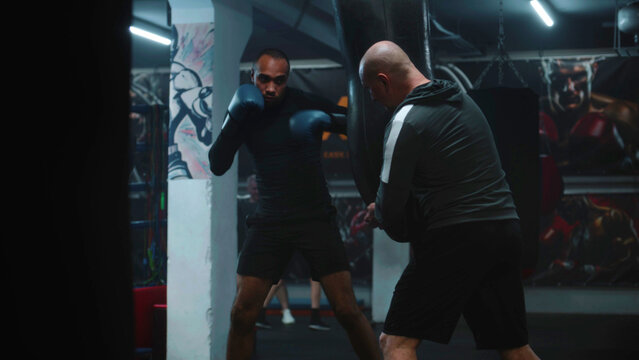 Confident African American fighter hits punching bag while training in dark boxing gym. Athletic man in boxing gloves exercises with coach before tournament. Physical activity and intensive workout.