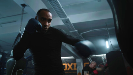 African American fighter listens coach command and starts hitting punching bag in dark boxing gym. Athletic man in boxing gloves exercises before tournament. Physical activity and intensive workout.