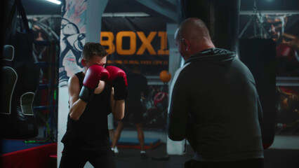 Young boxer in boxing gloves exercises with trainer in dark gym before fighting competition....