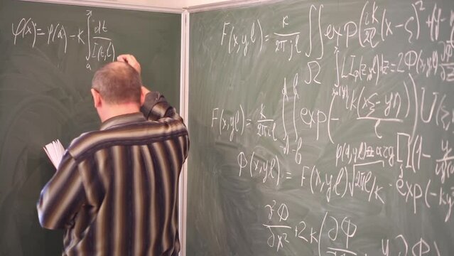 Teacher copying integral equations from book to blackboard in classroom.