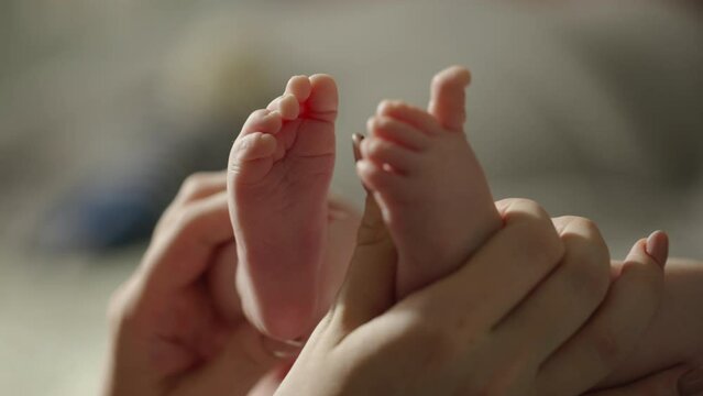 Woman hands hold baby's legs. Small tender feet of baby, hands of mother stroke baby's feet. Sun light shines on baby's legs and mommy mother hands. Close up. Concept of tenderness, love, affection.