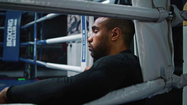 Tired and exhausted African American boxer sits in boxing ring corner. Fighter with beads of sweat on face rests after training. Athlete prepares to tournament or competition in boxing gym. Close up.
