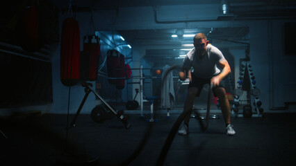 Male athlete exercises with battle ropes in dark boxing gym with LED lighting. Professional boxer does cardio or endurance workout before championship fight. Physical activity and CrossFit training.