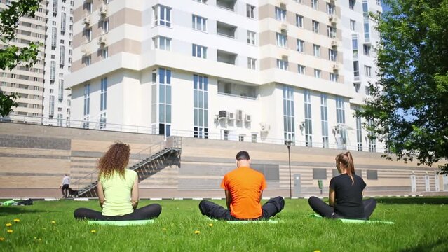 Back of man and two women on lawn sit and wave legs in butterfly pose of yoga