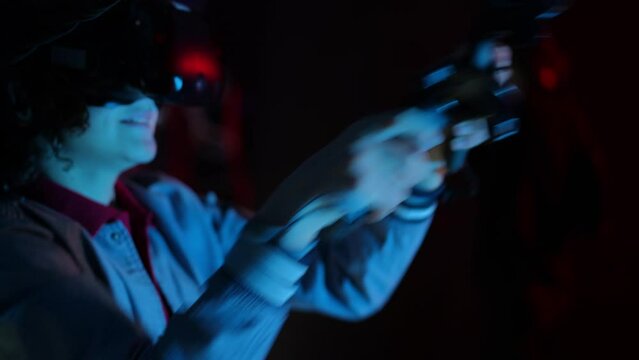 Close-up. A teenager wearing VR glasses and with controllers in his hands moves his hands quickly while standing in the dark of a VR club with blue lighting