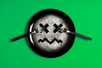 Sad face, face I don't like, concept made with plate and flour, green background, x-shaped eyes, black plate, sad mood, facial expressions, forks in the shape of arms with different positions